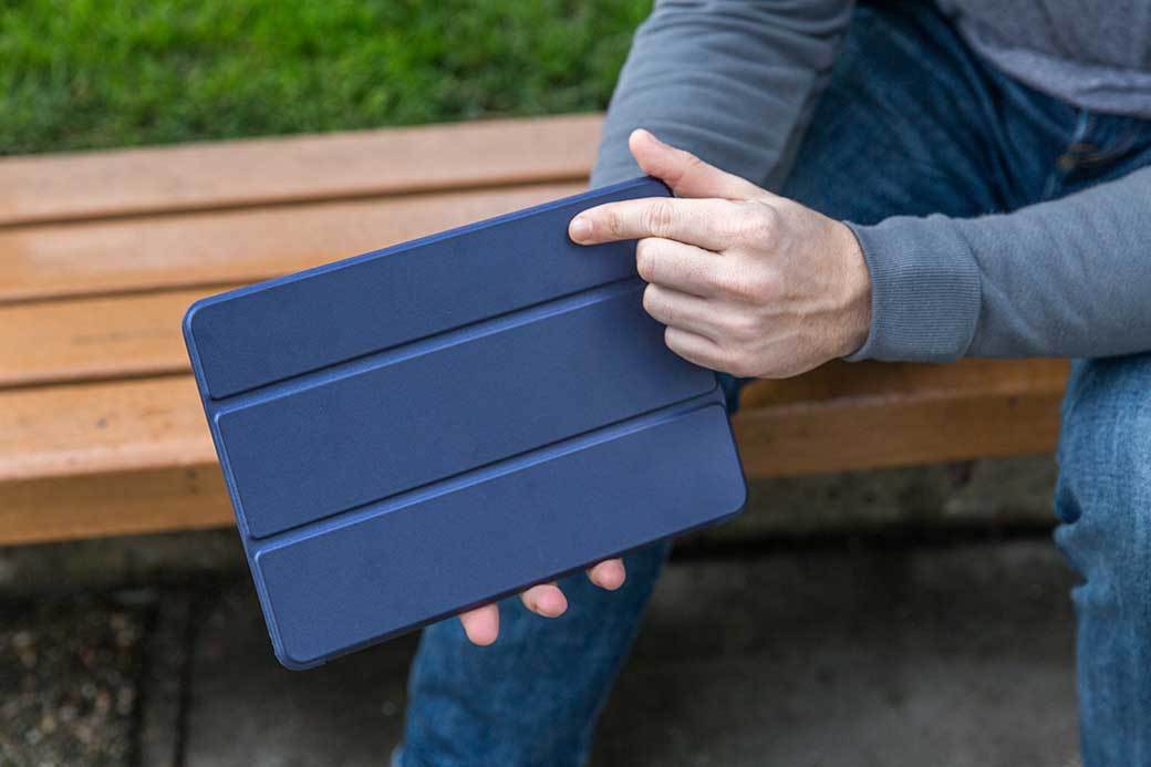 A person in a long sleeved blue shirt holds a tablet with a blue cover while sitting on a park bendch.