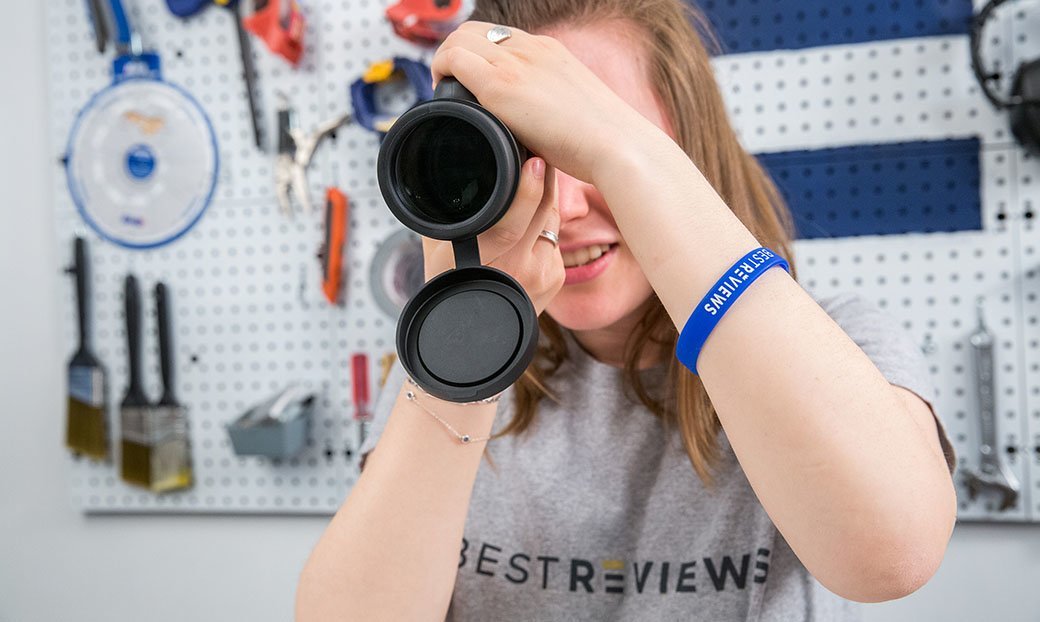 A long haired person standing in front of a wall of tools tests out a monocular